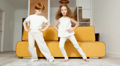 Two children standing with hands on hips in front of a yellow sofa