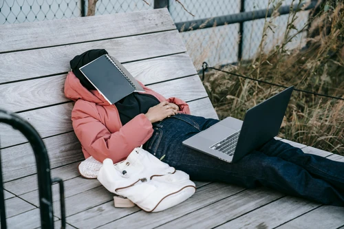 Tired woman napping on outdoor wooden lounge with notebook covering her face and laptop on lap