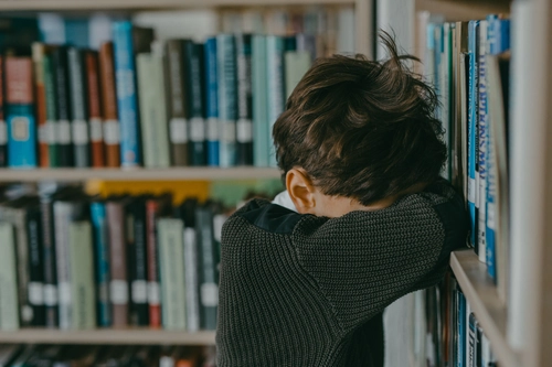 Boy buries his head between bookshelves, overwhelmed by school anxiety.