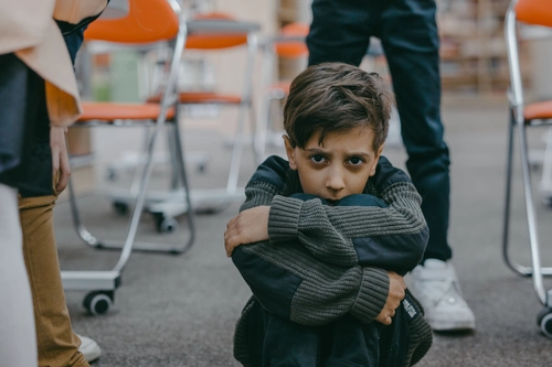 Anxious little boy hugs his knees tightly, surrounded by adults at school